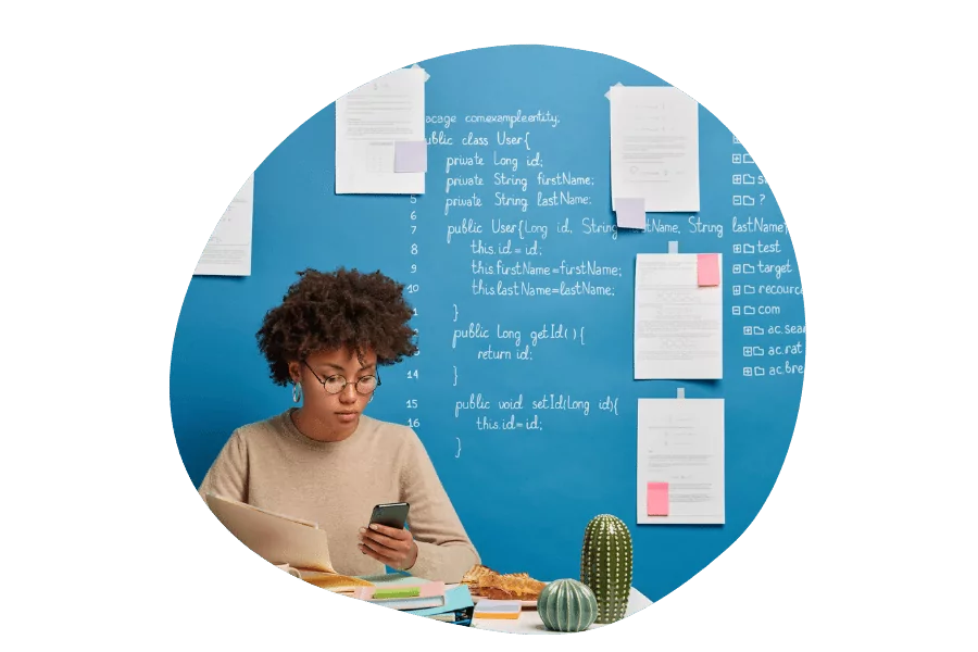 Femme assise à un bureau avec un ordinateur portable et un smartphone, devant un mur bleu couvert de feuilles et de lignes de code.